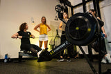 Person using a Concept2 rowing machine with others watching in a gym setting